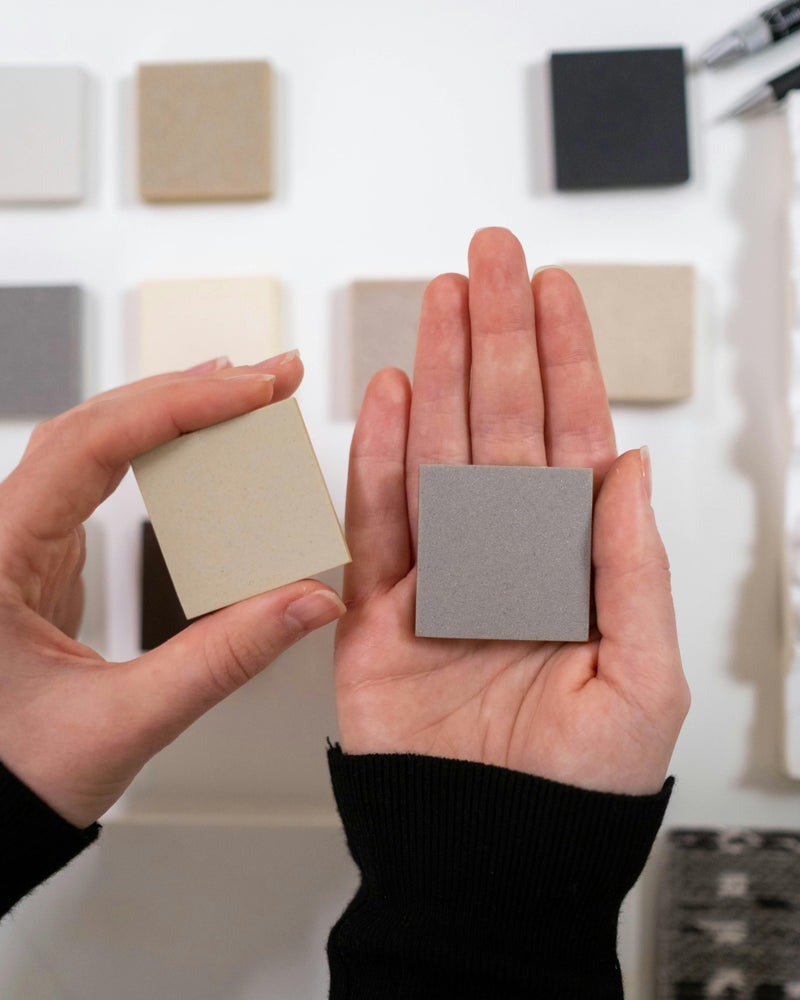 Person holding two small square samples of tile against a wall with more swatches.