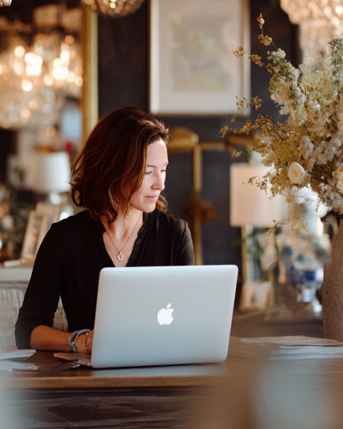 Woman using a laptop in a stylishly decorated room with chandeliers and a vase of flowers.